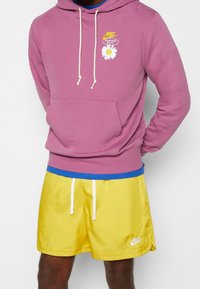 Růžová mikina Nike s bílými šňůrkami a grafikou sedmikrásky, v kombinaci se žlutými saténovými šortkami s bílými akcenty a logem.