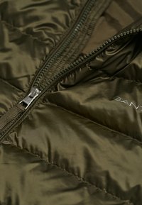 Chaqueta puffer de color verde olivo con un exterior brillante, que cuenta con un cierre frontal y una textura acolchada en forma de diamante. El logotipo de la marca se muestra de manera sutil.
