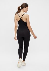Mono ajustado negro con escote en la espalda, tirantes delgados y textura sin costuras; las mallas se estrechan hasta el tobillo, combinadas con zapatillas blancas.