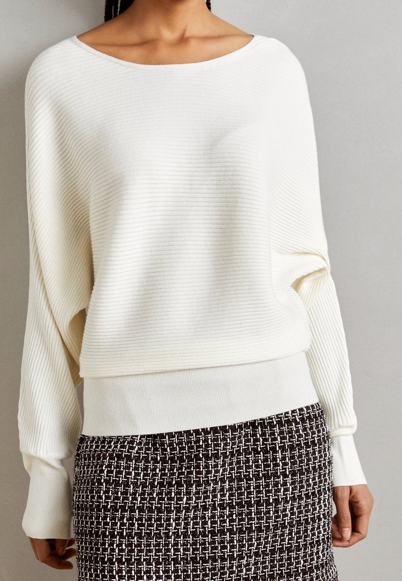 Femme portant un pull en maille côtelée blanche à manches longues et une jupe en tweed noir et blanc sur un fond gris uni.
