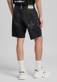 Shorts di denim nero con orlo grezzo, caratterizzati da un sottile motivo a croce sulla gamba e una patch brandizzata sul retro. Abbinati a scarpe sportive bianche e calze.