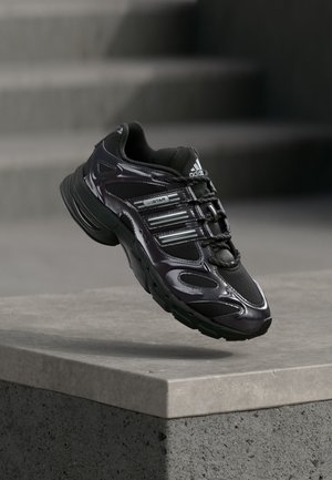 Czarny sneaker Adidas z szarymi paskami unoszący się nad szarą betonową powierzchnią i schodami w tle.