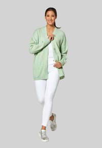 Lichtgroene geribbelde cardigan met een v-hals, twee voorzakken en knopen. Gecombineerd met een wit topje en witte aansluitende broek, en zilveren sneakers.