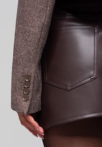 Manica di un blazer a spina di pesce marrone, con dettaglio del bottone, abbinata a una mini gonna in pelle marrone con tasca posteriore e texture liscia.