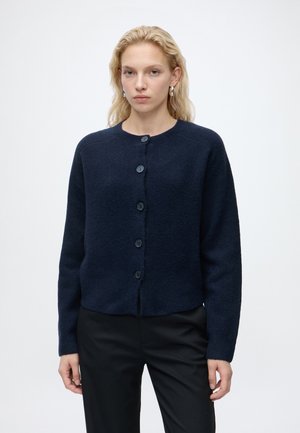 Femme blonde portant des boucles d'oreilles en argent, un cardigan bleu foncé boutonné et un pantalon noir, debout devant un fond blanc uni.