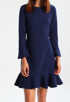 Femme portant une robe bleu marine à manches longues avec des poignets évasés et un ourlet à volants, debout devant un fond blanc uni.