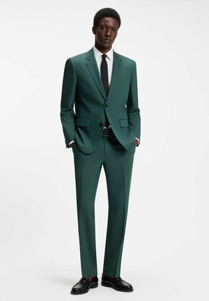 Młody mężczyzna stoi, ubrany w dopasowany ciemnozielony garnitur, białą koszulę, czarny krawat i czarne eleganckie buty, z rękami w kieszeniach.