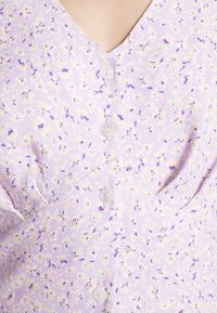 Blusa de tecido lilás com decote em V, apresentando um padrão floral em roxo, verde e tons pastel. Seis botões visíveis na parte da frente.