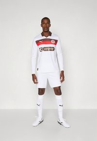 Hvit langermet fotballdrakt med brune og røde detaljer, med en logo på brystet, kombinert med hvite shorts og sokker.