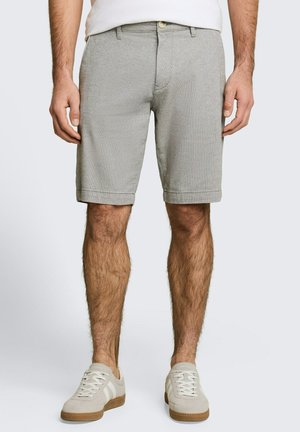 Homme portant un short gris clair longueur genoux, des baskets blanches et une chemise blanche, debout devant un fond clair uni.