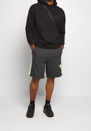 Homme portant un sweat à capuche noir, un short de sport gris foncé avec des accents néon, des chaussettes noires et des baskets noires, debout les mains dans les poches.