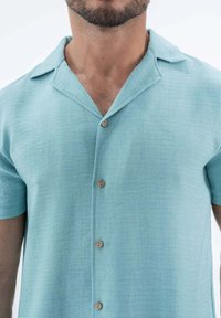 Chemise courte à manches en bleu clair, fabriquée en tissu texturé, avec un col peu profond et cinq boutons en bois sur le devant.