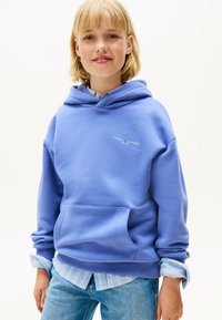 Jonge persoon met blond haar die een blauwe Tommy Hilfiger-hoodie draagt over een gestreept shirt en lichtblauwe jeans, met een lichte glimlach, op een witte achtergrond.
