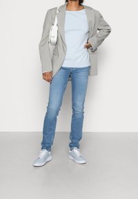 Person in einem hellgrauen Blazer, blassblauen Pullover, blauen Jeans, hellblauen Sneakern und mit einer weißen Umhängetasche, der auf einem grauen Boden steht.
