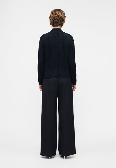 Άτομο με κοντά, σγουρά μαλλιά φοράει μαύρο πουλόβερ πλεκτό με τύπο καλωδίου και μαύρο παντελόνι με φαρδιά πόδια, στέκεται γυρισμένο προς τα πίσω σε λευκό φόντο.