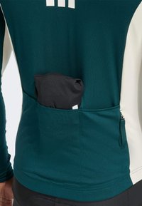 Long-sleeve athletic shirt in dark teal with a white accent. Features a rear pocket holding a folded black item and a zippered side pocket.