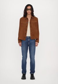 Brun ruskindsjakke med to brystlommer, beige top under, blå denim jeans og sorte støvler. Model iført grønne solbriller.