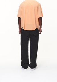 T-shirt met korte mouwen in perzikkleur met een losse pasvorm, gecombineerd met zwarte wijde broek en witte sneakers, gezien van achteren.