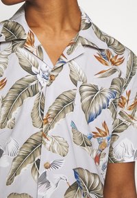 Uomo che indossa una camicia a maniche corte, con bottoni marroni, caratterizzata da un motivo tropicale di foglie e uccelli beige, bianche e blu su un tessuto chiaro.