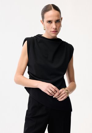 Frau mit zurückgekämmtem Haar, die ein ärmelloses schwarzes Oberteil und schwarze Hosen trägt, dazu goldene Ohrringe und ein massives goldenes Armband als Accessoires.