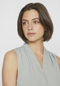 Helles graues ärmelloses Blusenshirt mit V-Ausschnitt, das einen weichen Stoff und dezente Falten aufweist. Keine sichtbaren Muster oder hardware Details.