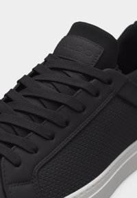 Baskets basses noires avec panneau latéral texturé, lacets noirs assortis et "ALDO" embossé sur la languette, avec une semelle blanche.