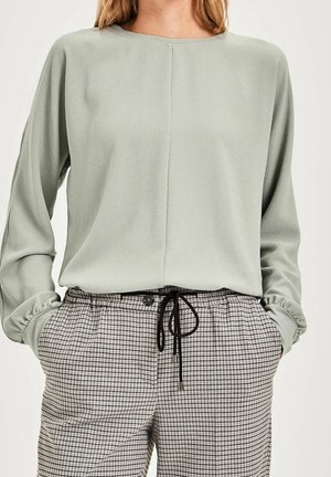 Personne portant un haut à manches longues vert sauge clair et un pantalon taille haute beige et noir à carreaux avec un cordon noir, les mains dans les poches.