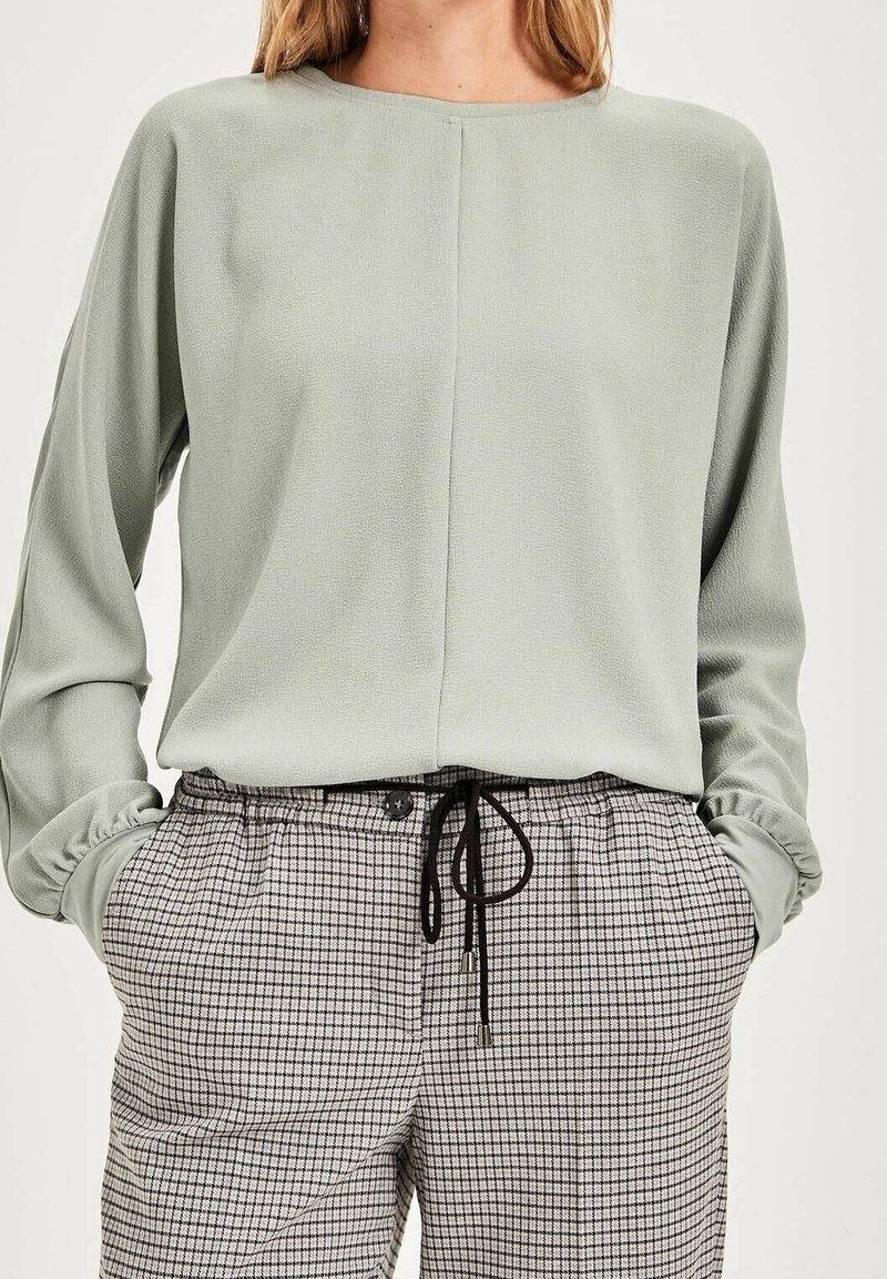 Personne portant un haut à manches longues vert sauge clair et un pantalon taille haute beige et noir à carreaux avec un cordon noir, les mains dans les poches.