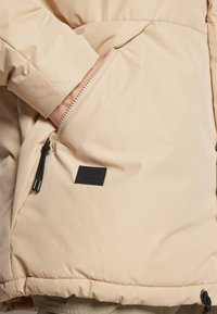 Beige puffjakke med glidelåslomme på siden, svart metall, og teksturert stoff. Merkevaren er tydelig på lommen. Glatt overflate.