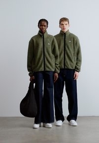 Grønne fleecetrøjer med sorte lynlåse, kombineret med mørke denimbukser. Personen til venstre holder en sort taske og bærer hvide sko.