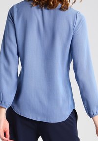 Světle modrá pruhovaná halenka se třičtvrtinovými rukávy, mírně zaobleným lemem a hladkou strukturou. Obsahuje vertikální bílé pruhy po celé ploše.