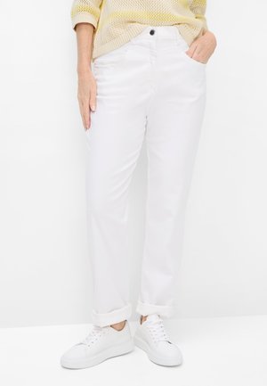 Femme portant un pantalon blanc retroussé, des baskets blanches et un pull en maille jaune clair, une main dans la poche.