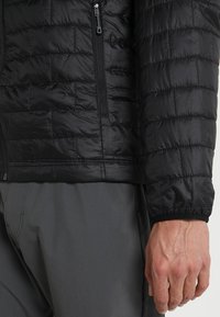 Svart vadderad jacka med quiltad textur, utrustad med en dragkedjeförsedd ficka och elastiska muddar. Bärs över gråa byxor, visar närbild på ärmen.