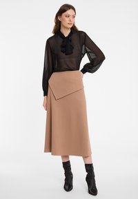 Schwarze transparente Bluse mit einer Schleife, kombiniert mit einem beigen A-Linien-Rock mit asymmetrischem Faltenwurf. Das Model trägt schwarze Stiefeletten.