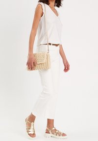 Beige woven crossbody bag with a flap, featuring a textured finish. Worn with white jeans and a light top, complemented by metallic sandals.