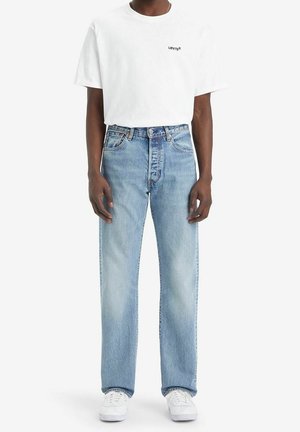 Man draagt een wit Levi's T-shirt, lichtblauwe recht gesneden jeans en witte sneakers, staand tegen een effen achtergrond.