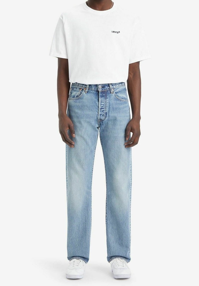 Homme portant un t-shirt blanc Levi's, un jean bleu clair à jambes droites et des baskets blanches, debout devant un fond uni.