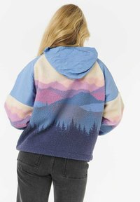 Blond kvinde iført en flerfarvet fleecejakke med et bjerg- og skovmønster, blå hætte og sorte jeans, vendt væk.