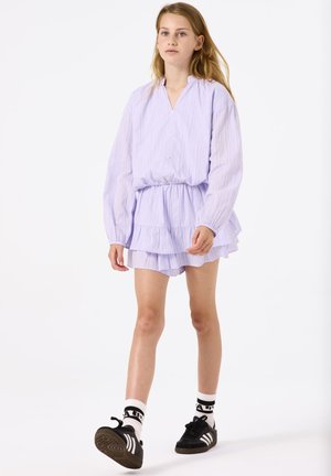 Helllila geschichtetes Kleid mit langen Ärmeln, Falten und einem gerafften Taillenbund, kombiniert mit schwarzen Sportschuhen und gemusterten Socken.