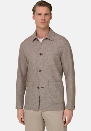 Uomo che indossa una giacca beige testurizzata con bottoni e tre tasche sopra una camicia bianca e pantaloni chiari, in piedi davanti a uno sfondo semplice.