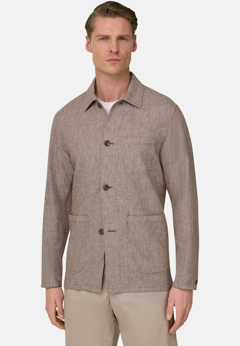 Homme portant une veste beige texturée à boutons avec trois poches, sur une chemise blanche et un pantalon clair, se tenant devant un fond uni.