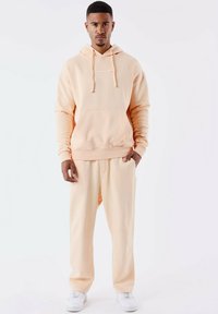 Sudadera de color durazno con bolsillo frontal, capucha con cordones y pantalones de chándal a juego. Textura suave, corte relajado, combinada con zapatillas blancas.