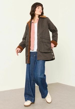 Frau trägt eine braune Jacke mit Lammfellkragen, rosa Hemd, weite blaue Jeans und weiße Schuhe, steht und schaut zur Seite.
