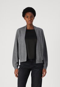 Cardigan de punto gris con un ajuste relajado, delantero abierto y mangas largas. Llevado sobre una parte superior negra, combinado con jeans oscuros. Textura suave.