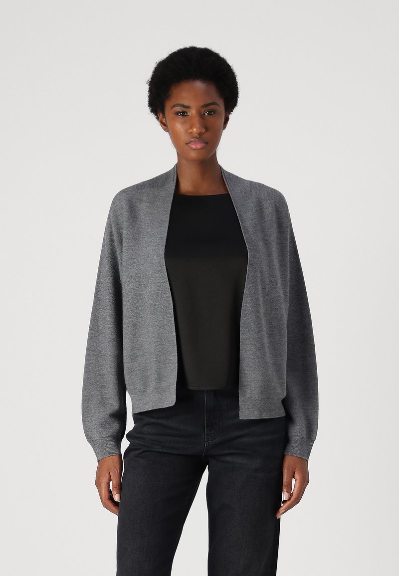 Cardigan de punto gris con un ajuste relajado, delantero abierto y mangas largas. Llevado sobre una parte superior negra, combinado con jeans oscuros. Textura suave.
