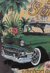 Coche vintage verde con acentos de cromo y un detallado adorno en el capó, situado frente a un fondo de palmeras y flores sobre un fondo texturizado amarillo.