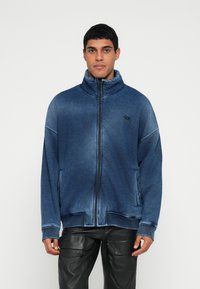 Modra jopa z zadrgo in visokim ovratnikom, izdelana iz mehke tkanine. Ima rebraste manšete in spodnji rob, dva stranska žepa ter logotip na prsih.