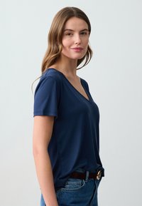 Niebieska koszulka z krótkim rękawem i dekoltem w serek, wykonana z miękkiego materiału, zestawiona z wysokim stanem dżinsami i czarnym paskiem z złotą klamrą.