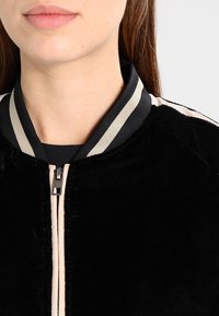 Giubbotto bomber nero con una texture morbida, dotato di colletto a costine a righe beige e nere, e chiusura con zip.