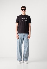 Tricou negru din bumbac cu "CALVIN KLEIN" imprimat în alb, purtat cu blugi lejeri de culoare bleu deschis și adidași albi, model purtând ochelari de soare.